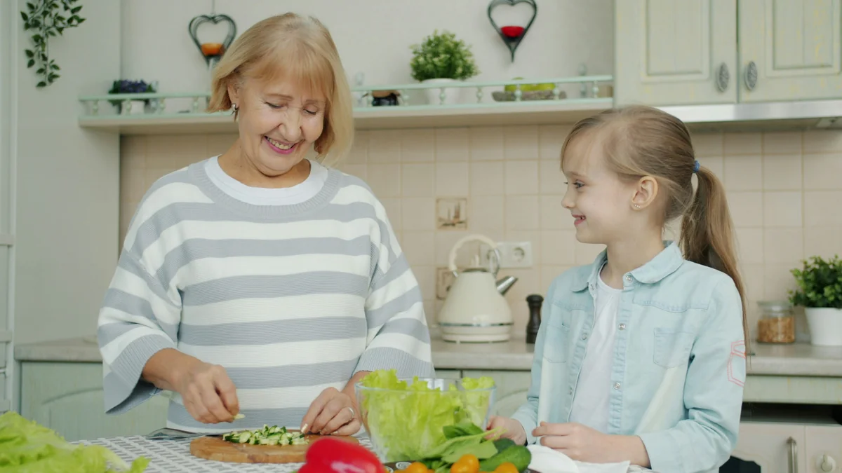 Senior grandmother is making salad and feeding cute happy child with vegetable chops expressing love having fun in kitchen at home. People and lifestyle concept. - family learning activities