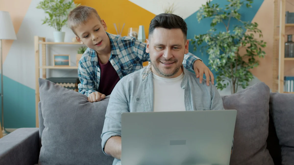 Happy child coming to father working with laptop at home talking using computer together in cozy living room. Communication and modern gadgets concept. - family learning activities