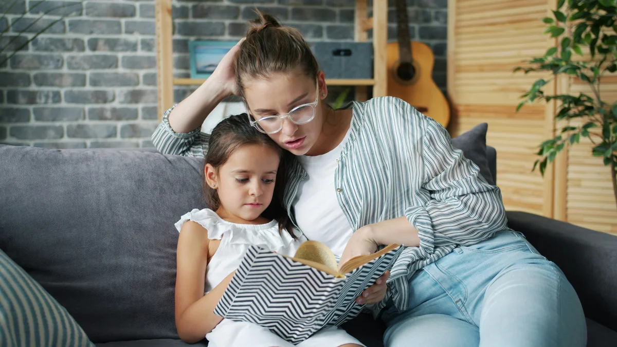 Happy young family mother and daughter are reading children's book at home smiling enjoying time together. Apartment, culture and childhood concept. - family learning activities