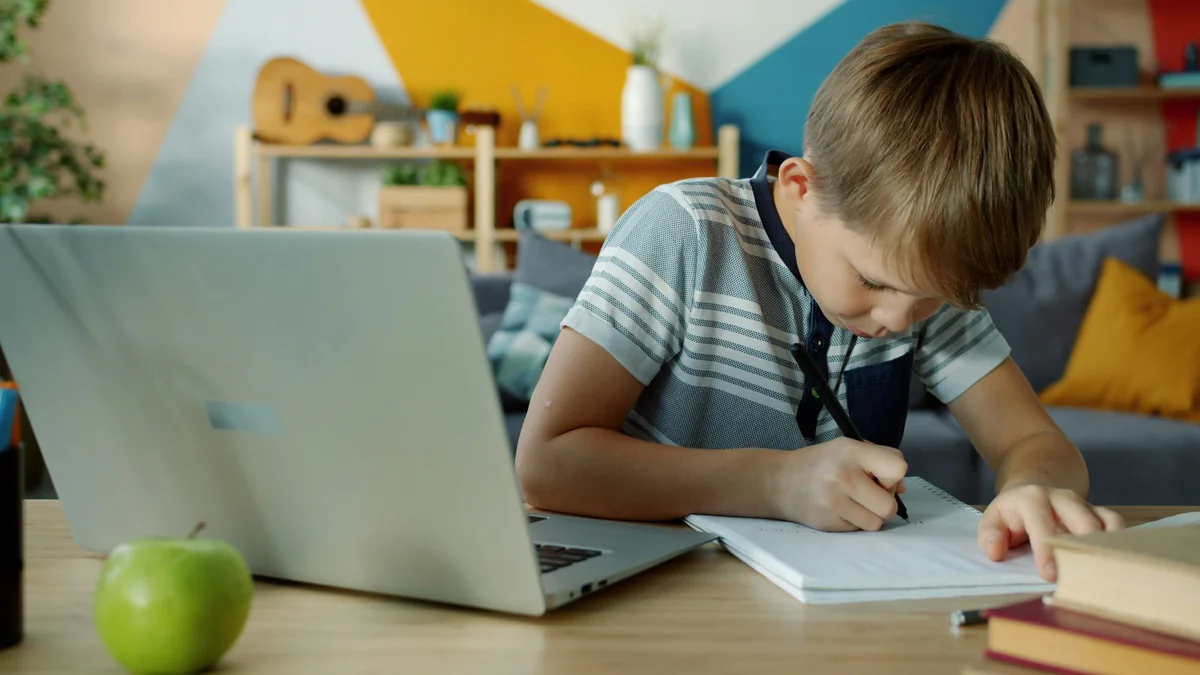 happy child using laptop for education writing in notebook doing homework sitting at desk in apartment. Technology and lifestyle concept. - home schooling strategies