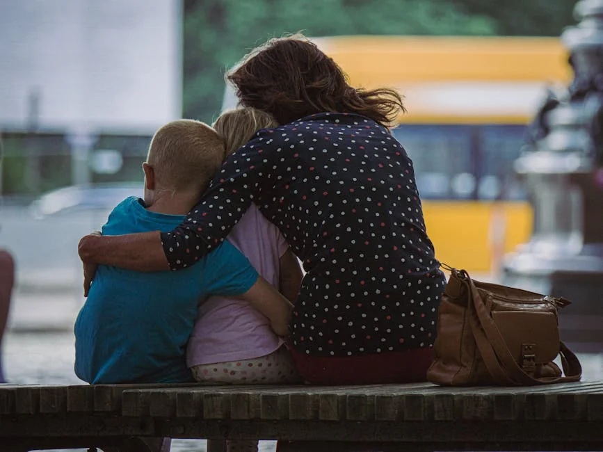 A mother hugging her two children while sitting on a park bench outdoors. - family children care - Parenting tips